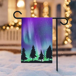 May include: A garden flag featuring a vibrant depiction of the Aurora Borealis. The design showcases a purple and green sky with stars, silhouetted pine trees, and a black metal stand. The flag is displayed outdoors in a snowy setting.