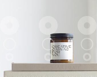 May include: A brown glass candle jar with a black lid and a white label that reads "CREATIVE CANVAS FOR YOU". The candle is sitting on top of two books.