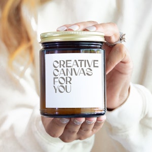 Puede incluir: Un tarro de vidrio marrón con tapa dorada, sostenido en las manos. El tarro tiene una etiqueta blanca con el texto "CREATIVE CANVAS FOR YOU" en gris. El tarro parece ser una vela o un producto similar.