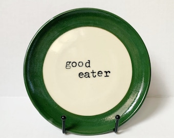 May include: A white ceramic plate with a green rim and the words "good eater" printed in black.