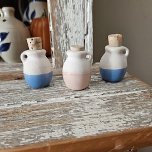 May include: Three small, ceramic bottles with cork stoppers. Each bottle is white on top and has a colored base: blue, pink, and blue. The bottles have small handles and are displayed on a distressed wooden surface.