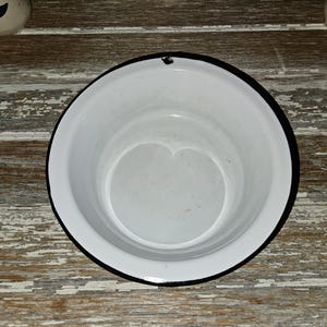 May include: A white enamel bowl with a black rim, set against a weathered, distressed wooden surface. The bowl has a simple, classic design, and is empty. The background includes glimpses of other ceramic items.