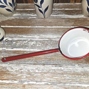 May include: A vintage red and white enamelware ladle with a long handle. The ladle has a white interior and a red rim and handle. The handle has a hole for hanging. The ladle is placed on a weathered wooden surface.