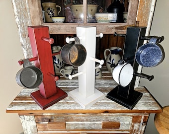 May include: Three wooden mug trees in red, white, and black, each featuring a square central post and a square base. The red and white stands are empty, while the black stand holds two small metal mugs with speckled finishes.