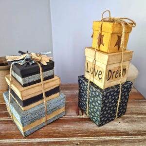 May include: Two stacks of square decorative boxes tied with twine. The left stack features black, tan, and blue patterned boxes with frayed fabric ribbon. The right stack includes a yellow box with stars and a tan box with the text Love Dream.