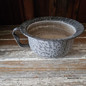 May include: A gray and white speckled enamelware bowl with a single handle. The bowl is sitting on a weathered wooden surface.