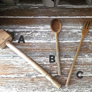 May include: A wooden meat mallet, spoon, and fork rest on a distressed, white-painted wooden surface. The kitchen utensils are made of light-colored wood. The mallet has a rectangular head. The spoon has a rounded bowl. The fork has four tines.