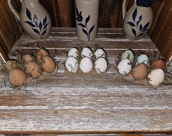 May include: Three sets of decorative eggs in egg cartons, with brown, white, and blue-green eggs. The eggs are nestled in straw and surrounded by dried flowers. The egg cartons are on a wooden surface.