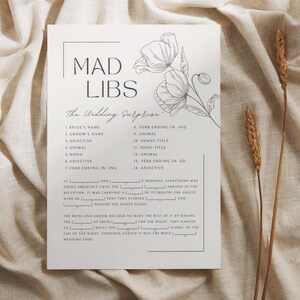 May include: A white Mad Libs wedding game card with a floral illustration. The card features fill-in-the-blank prompts for a wedding-themed story. The text includes "The Wedding Surprise" and numbered blanks for nouns, adjectives, and verbs.
