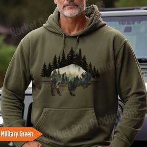 May include: A military green hooded sweatshirt featuring a graphic print of a bison silhouette filled with a forest scene, set against a background of black pine tree silhouettes. The hoodie includes a front kangaroo pocket and matching drawstrings.