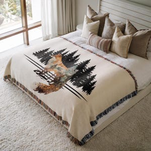 May include: A cream-colored woven throw blanket featuring a graphic print of a fox standing among black pine trees. The blanket has a fringed edge with multi-colored threads and is draped over a bed with several neutral-toned pillows.