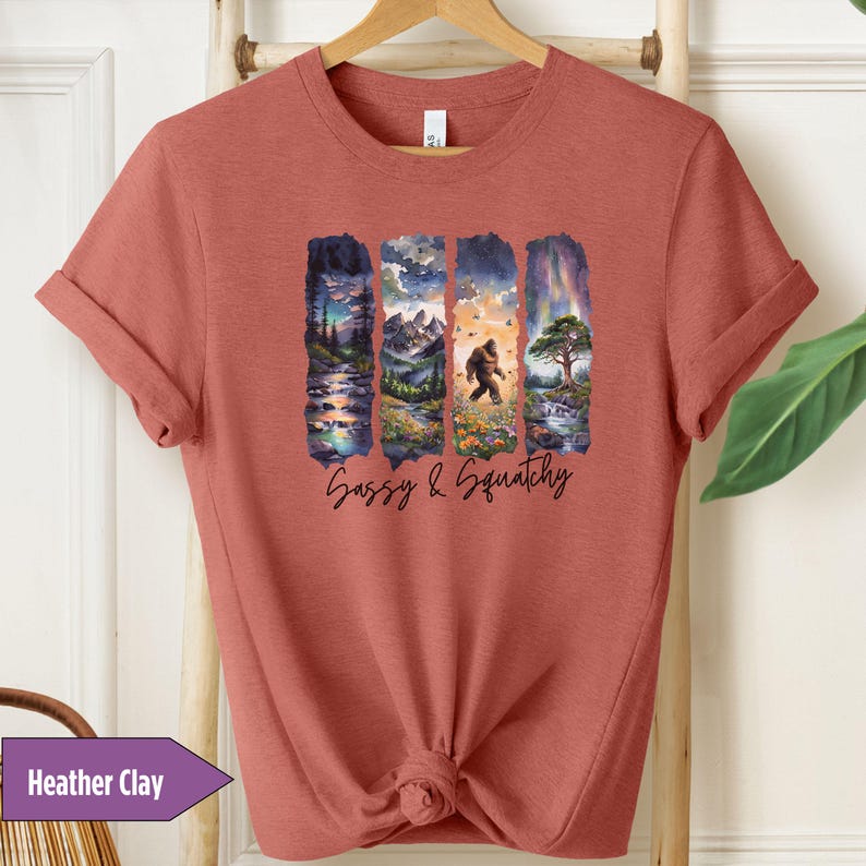 May include: A heather clay t-shirt with a graphic print of a sasquatch walking through a forest. The text "Sassy & Squatchy" is printed below the graphic.