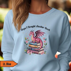 May include: Light blue sweatshirt featuring a pink dragon perched atop a stack of books. The dragon has purple wings and is surrounded by flowers. The text "Oops I Bought Another Book" is above the dragon.
