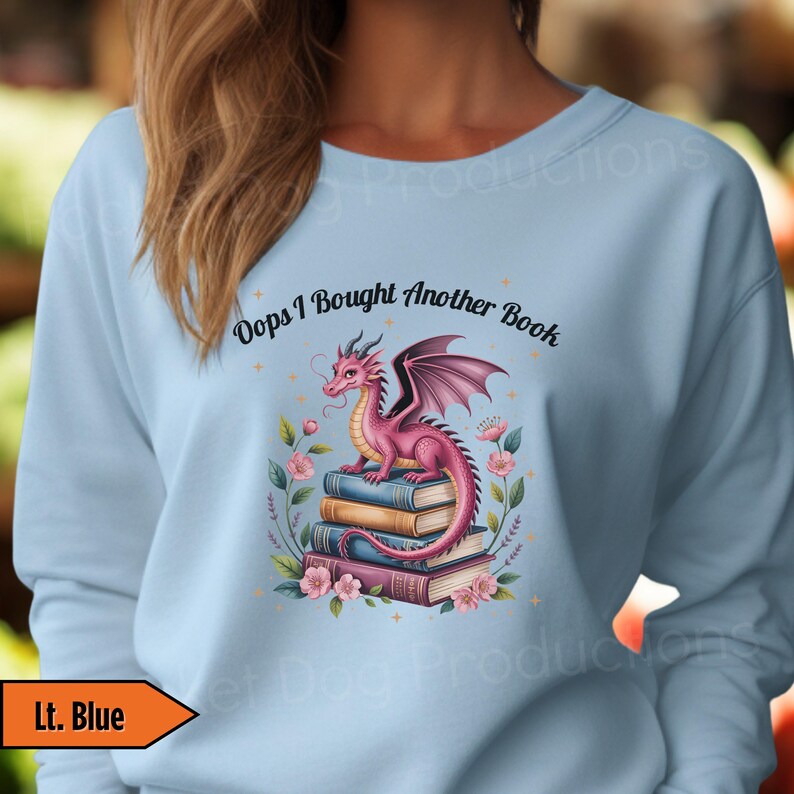 May include: Light blue sweatshirt featuring a pink dragon perched atop a stack of books. The dragon has purple wings and is surrounded by flowers. The text "Oops I Bought Another Book" is above the dragon.