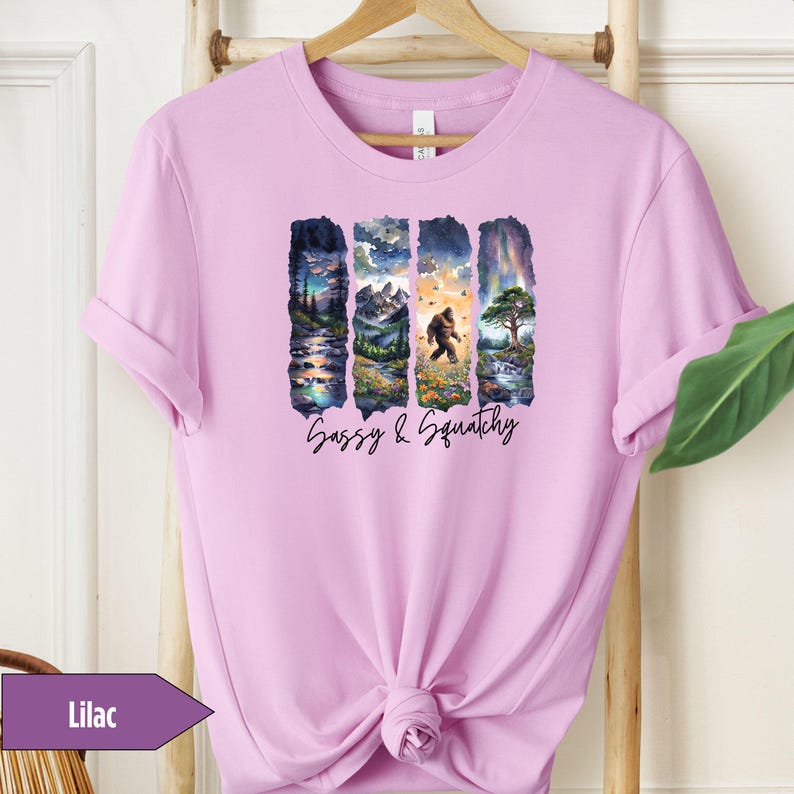 May include: Lilac t-shirt with a graphic print of four nature scenes with a sasquatch walking through one of the scenes. The text "Sassy & Squatchy" is printed below the graphic.