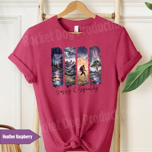 May include: A heather raspberry t-shirt with a graphic of a Bigfoot walking through a forest scene. The text "Sassy & Squatchy" is printed below the graphic.