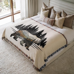 May include: A woven throw blanket featuring a large bear silhouette filled with a landscape of mountains and trees. The bear is set against a black forest graphic on a cream-colored background. The blanket has a fringed edge with multi-colored threads.