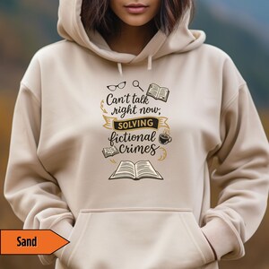 May include: Sand-colored hoodie with the text "Can't talk right now, solving fictional crimes" in black and gold. The design includes glasses, a magnifying glass, books, and a coffee cup. The hoodie has a drawstring hood and a front pocket.