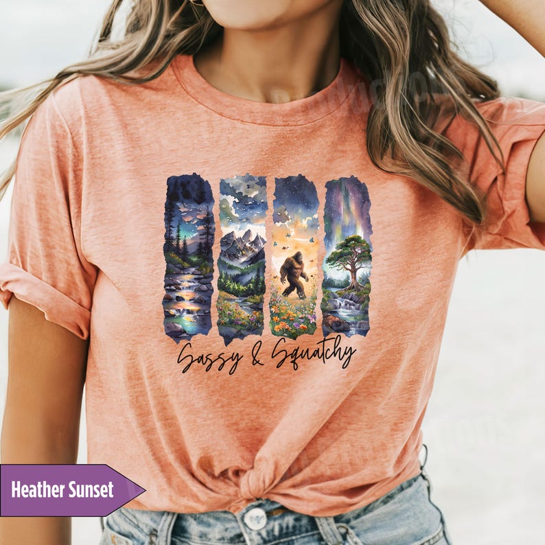 May include: A heather sunset colored t-shirt with a graphic print of four vertical watercolor scenes. The scenes include a mountain range, a forest, a waterfall, and a forest with a Bigfoot walking through it. The text 'Sassy & Squatchy' is printed below the scenes.