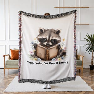 May include: A woven throw blanket featuring a central illustration of a raccoon reading an open book, surrounded by colorful pastel flowers. The text "Trash Pandas, But Make It Literary" is printed below the raccoon. The blanket has fringed edges.