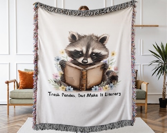 May include: A woven throw blanket featuring a central illustration of a raccoon reading an open book, surrounded by colorful pastel flowers. The text "Trash Pandas, But Make It Literary" is printed below the raccoon. The blanket has fringed edges.