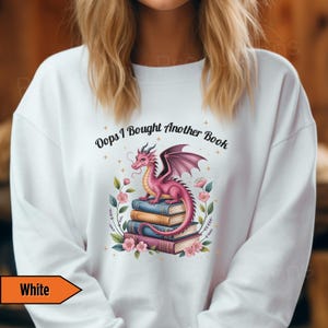 May include: White sweatshirt featuring a pink dragon perched atop a stack of books. The dragon has purple wings and is surrounded by flowers. The text "Oops I Bought Another Book" is arched above the dragon.