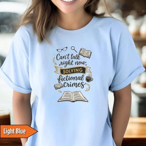 May include: Light blue t-shirt with the text "Can't talk right now, solving fictional crimes." The graphic includes glasses, a magnifying glass, books, and a coffee cup. The shirt is a light blue color.