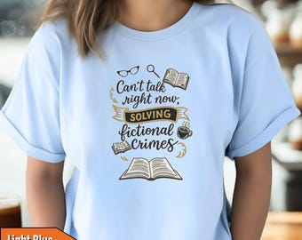 May include: Light blue t-shirt with the text "Can't talk right now, solving fictional crimes." The graphic includes glasses, a magnifying glass, books, and a coffee cup. The shirt is a light blue color.