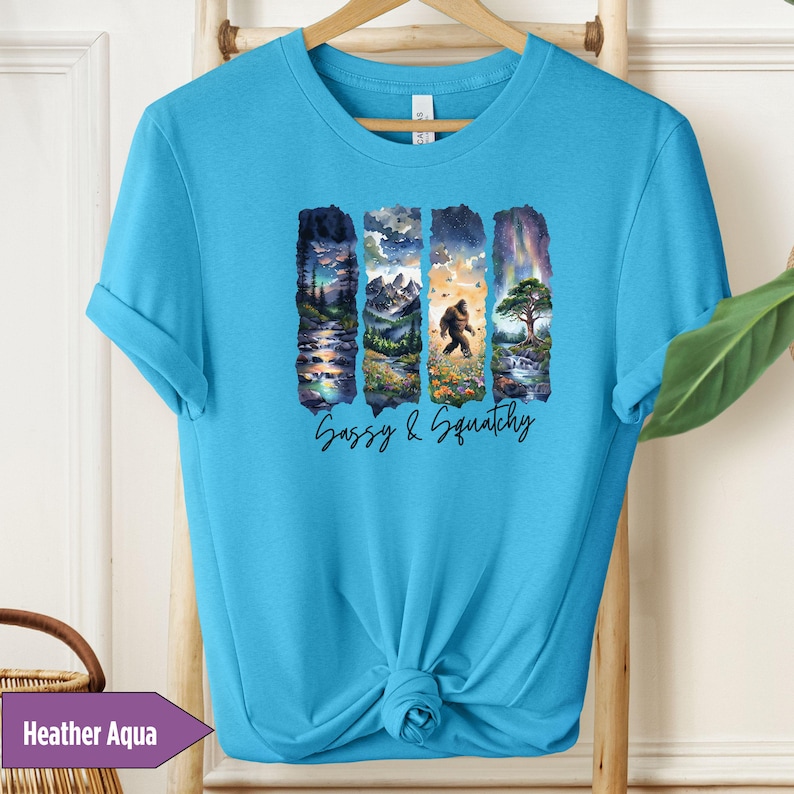 May include: A turquoise blue t-shirt with a graphic print of a sasquatch walking through a forest scene. The text "Sassy & Squatchy" is printed below the graphic.