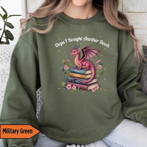 May include: Military green sweatshirt with the text "Oops I Bought Another Book." The graphic features a pink dragon sitting atop a stack of books, surrounded by flowers. The dragon has purple wings and is facing to the right.
