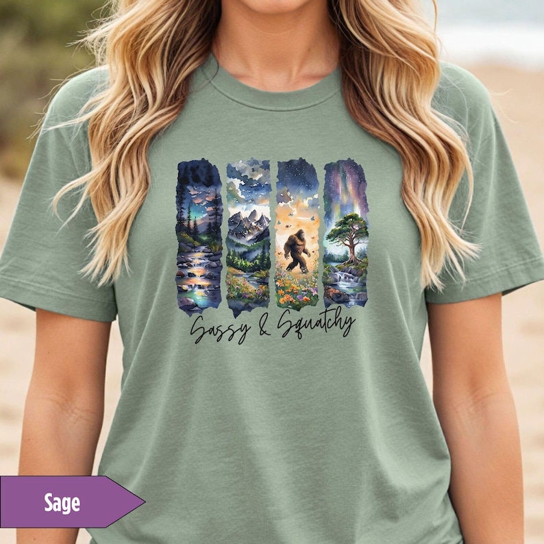 May include: Sage green t-shirt with a graphic print of four vertical panels featuring a nature scene with a sasquatch walking through a forest. The text 'Sassy & Squatchy' is printed below the graphic.