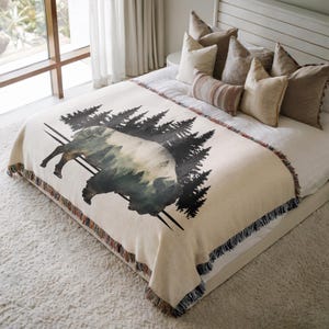 May include: A cream-colored woven throw blanket featuring a large graphic of a bison silhouette filled with a forest scene. The design includes a dark black pine tree forest background and a fringed edge along the bottom of the blanket.