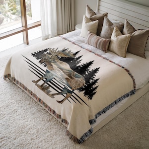 May include: A cream-colored woven throw blanket featuring a large graphic of a moose silhouette filled with a forest landscape scene. The design includes black pine tree silhouettes and horizontal lines, finished with a fringed edge on all sides.