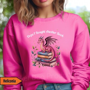 May include: Pink sweatshirt with a graphic of a pink dragon perched on a stack of books, surrounded by flowers. The text "Oops I Bought Another Book" is above the dragon. The word "Heliconia" is in a yellow box.