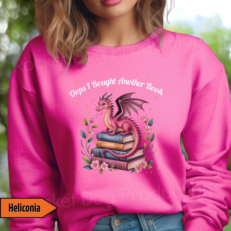 May include: Pink sweatshirt with a graphic of a pink dragon perched on a stack of books, surrounded by flowers. The text "Oops I Bought Another Book" is above the dragon. The word "Heliconia" is in a yellow box.