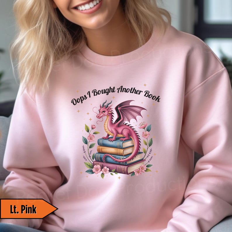 May include: Light pink sweatshirt with the text "Oops I Bought Another Book." The graphic features a pink dragon perched on a stack of books, surrounded by flowers. The dragon has dark purple wings and is facing left.