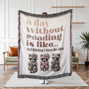 May include: A cream-colored woven throw blanket featuring a fringed edge and the brown text "a day WITHOUT reading is like... Just Kidding I Have No Idea". Below the text are three illustrated raccoons, each holding a book and wearing flowers.