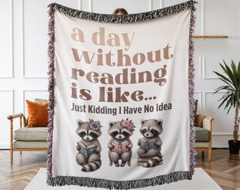 May include: A cream-colored woven throw blanket featuring a fringed edge and the brown text "a day WITHOUT reading is like... Just Kidding I Have No Idea". Below the text are three illustrated raccoons, each holding a book and wearing flowers.