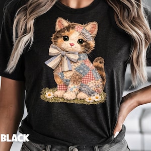 Op de afbeelding: Zwart t-shirt met een patchwork katje illustratie. Het katje heeft een strik en zit op gras met madeliefjes. Het woord "BLACK" staat onderaan het shirt.
