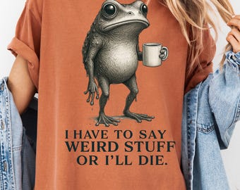 May include: Burnt orange t-shirt featuring a detailed illustration of a frog holding a mug. The frog has large eyes and a textured skin. The text below reads: "I HAVE TO SAY WEIRD STUFF OR I'LL DIE."