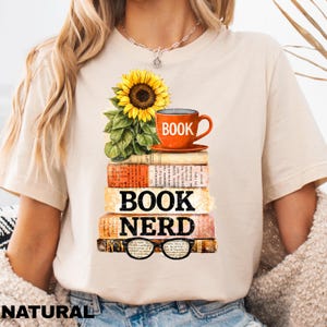 May include: Beige t-shirt with a graphic design featuring a stack of books, a sunflower, and an orange coffee cup. The words "BOOK NERD" are printed on the books, with eyeglasses at the bottom. The word "NATURAL" is printed at the bottom of the shirt.