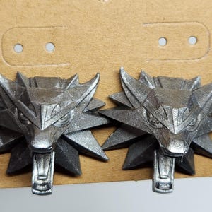 May include: A pair of silver-colored wolf head earrings. The wolf heads are detailed and have a sharp, menacing look. The earrings are a popular symbol of the Witcher video game series.