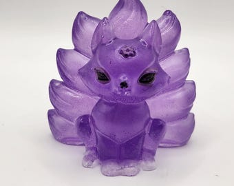 Puede incluir: Una figura de zorro morado con nueve colas. El zorro está sentado erguido con las patas delanteras. La figura tiene ojos negros y una nariz pequeña. Las colas están extendidas detrás del zorro. La figura está hecha de un material translúcido.