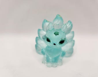 Peut inclure: Figurine de renard bleu clair avec plusieurs queues. Le renard a des yeux noirs et un petit nez. Les queues sont superposées et s'éventent derrière le renard. La figurine a un petit motif vert sur le front.