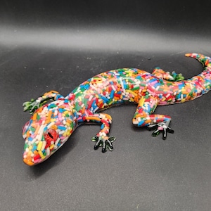 Puede incluir: Una colorida figura de lagarto cubierta con un patrón de chispas multicolores. El lagarto tiene ojos rojos, pies verdes y una cola curvada. La figura está sobre una superficie oscura.