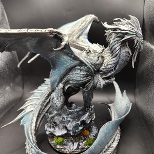 May include: A detailed figurine of a dragon with a silver and blue color scheme. The dragon is perched on a rocky base with green moss. Its wings are spread, and its eyes are green. The dragon's tail curves around the base.