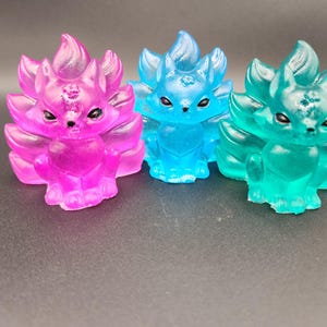 Puede incluir: Tres figuras translúcidas de zorro en rosa, azul y verde. Cada zorro tiene múltiples colas y una cara detallada con ojos negros. Las figuras se exhiben sobre una superficie oscura, mostrando sus colores vibrantes y su intrincado diseño.