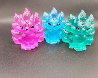 Puede incluir: Tres figuras translúcidas de zorro en rosa, azul y verde. Cada zorro tiene múltiples colas y una cara detallada con ojos negros. Las figuras se exhiben sobre una superficie oscura, mostrando sus colores vibrantes y su intrincado diseño.