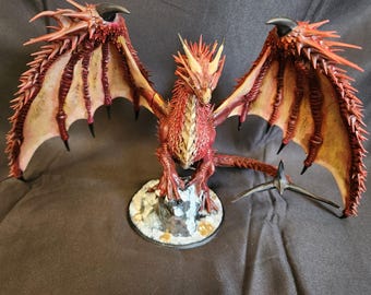 Può includere: Statuetta di drago rosso e giallo dettagliata con ali spiegate. Il drago ha artigli affilati, corna e una lunga coda. È montato su una piccola base rocciosa, le ali sono spalancate.
