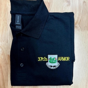 May include: A black polo shirt with a collar and three buttons. The chest features an embroidered emblem with a green dragon on a shield, flanked by the yellow text 37th and ARMOR, with a banner below reading COURAGE CONQUERS.